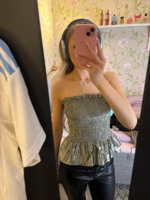 Glittrig silver tubtopp från & Other Stories - Säljer en glittrig silver tubtopp från & Other Stories, modell Stockholm Atelier. Toppen är bandeau med smockad överdel och volang nertill. Perfekt för dig som vill sticka ut med en metallic look. Superfin att styla med både jeans och kjol!