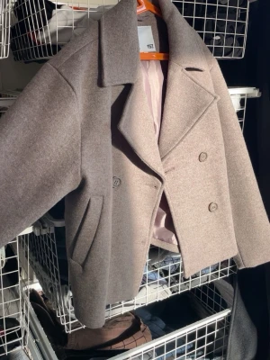 Beige kort skepparkavaj i ullmix - Kort skepparkavaj i beige ullmix från 157. Jackan har dubbelknäppning med stora knappar, bred krage och två snedställda fickor framtill. Fodrad med ljusrosa tyg på insidan. Perfekt för dig som vill ha en stilren och klassisk look. Passar mig som bär XS 