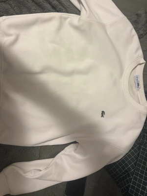 Vit sweatshirt från Lacoste, storlek L - Säljer en stilren vit sweatshirt från Lacoste i storlek L. Tröjan har rund hals, ribbade muddar och det klassiska gröna krokodilmärket broderat på bröstet. Mjuk insida och enkel design som funkar till många olika looks.