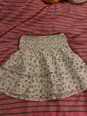 blommig volangkjol - Säker en supersöt kort kjol i vitt med små blommor i blått, grönt och rosa. Kjolen har bred smock i midjan och dubbla volanger som ger en flowy känsla. Perfekt för sommaren💐