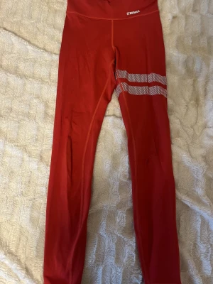 Röda leggings från Stronger - Säljer ett par röda leggings från Stronger med vita prickiga ränder på ena benet. De har hög midja och är gjorda i stretchigt syntetmaterial som passar perfekt för träning. Snygg och sportig design med logga framtill i vitt.
