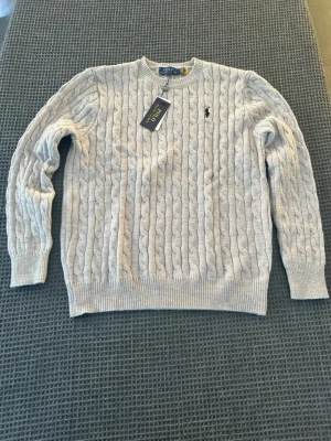Grå stickad tröja från Polo Ralph Lauren - Snygg grå helt ny stickad tröja från Polo Ralph Lauren med klassiskt kabelstickat mönster och rund hals. Tröjan har långärmade ribbade muddar och en broderad svart logga på bröstet. Perfekt för dig som gillar stilrena och tidlösa plagg. Tröjan är i storleken L men passar mer som M