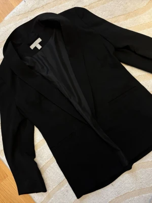 Svart kavaj från H&M, storlek 36 - Stilren svart kavaj från H&M i storlek 36. Klassisk modell med smal krage och två fickor framtill. Materialet är mjukt och har en lätt glansig insida. Perfekt för dig som gillar en clean och enkel look.