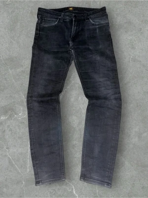 Svarta slim  jeans från Lee - Snygga svarta jeans från Lee med slim passform och klassisk femficksdesign. Jeansen har mörk tvätt med subtila slitningar och Lee-logga på bakfickan samt läderpatch i midjan. Perfekta för dig som gillar en smal siluett och stilren look.