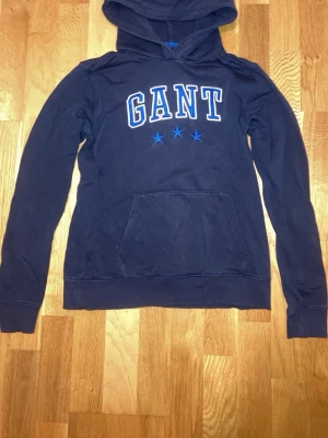 Mörkblå GANT hoodie. - Mörkblå GANT hoodie med luva och stilrent ljusblått tryck framtill. Klassisk och tidlös modell i mjukt och bekvämt material. Hoodien är i fint skick och passar perfekt till både vardag och avslappnade outfits. Ett måste för dig som gillar en clean och preppy stil. (Ej samma plagg som andra inlägget bara ett likadant).