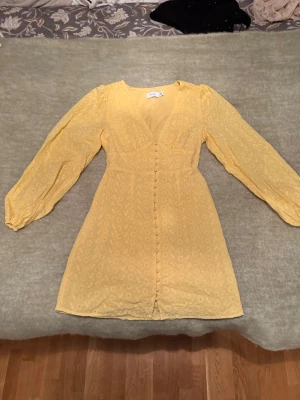 Gul kort klänning från Vila - Söt gul klänning från Vila med broderade blommor, v-ringad hals och långa puffärmar. Klänningen har knappar framtill och är tillverkad i ett lätt bomullstyg som känns luftigt. Perfekt för sommarens alla tillfällen!