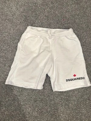 Vita shorts från DSQUARED2 med snörning - Säljer ett par vita shorts från DSQUARED2 med snörning i midjan och tryckt logga med röd lönn på ena benet. De har en avslappnad passform och är gjorda i mjukt bomullsmaterial.
