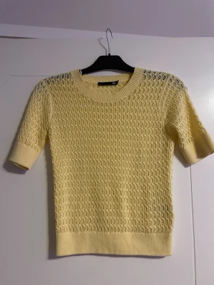 Ljusgul stickad tröja från 157 - Snygg ljusgul stickad tröja från 157 i storlek XS. Tröjan har ett unikt hålmönster, rund hals och ribbade muddar vid ärmslut och nederkant. Ärmarna är korta och materialet känns mjukt och luftigt, perfekt för vår och sommar.