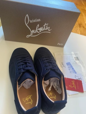 Mörkblå sneakers från Christian Louboutin - Säljer ett par mörkblå sneakers från Christian Louboutin med ikonisk röd sula och broderad logga på sidan och hälen. Skorna har snörning, rund tå och är tillverkade i mocka med beige innersula. Kommer med originalkartong och certifikat.