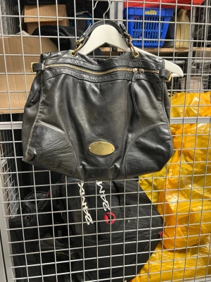 Svart axelväska från Mulberry i skinn - Säljer en svart axelväska från Mulberry i äkta skinn med guldfärgade metalldetaljer och dragkedja upptill. Väskan har en klassisk Mulberry-logga i metall framtill och ett rymligt huvudfack. Perfekt för dig som gillar stilrena och tidlösa accessoarer.