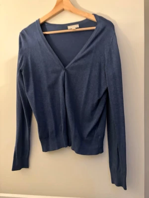 Blå kofta från H&M med v-ringning - Snygg blå kofta från H&M i storlek M. Koftan har en djup v-ringning och långa ärmar, perfekt att slänga på sig när det är lite kyligt. Materialet är mjukt och tunt, vilket gör den skön att bära under höst och vår.