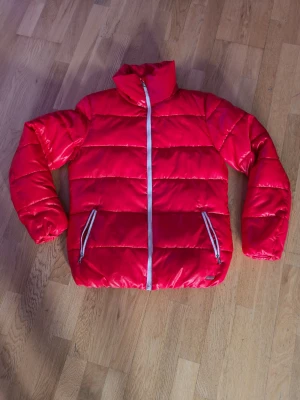 Röd pufferjacka från On The Peak - Riktigt snygg röd pufferjacka från On The Peak med vitt dragkedjor och hög krage. Jackan har två fickor med dragkedja och quiltad design som ger en cool look. Perfekt för kalla dagar när du vill sticka ut.
