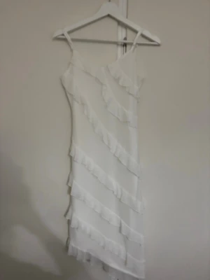 Vit långklänning med volanger - Säljer en vit långklänning med tunna axelband och flera diagonala volangdetaljer. Klänningen har en lätt och flödande form som ger en romantisk vibe. Perfekt för dig som gillar en feminin och stilren look.