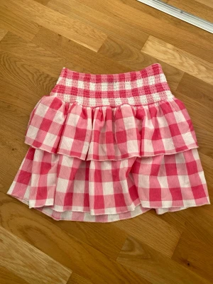 Kort rosa och vit volangkjol - Säljer en kort kjol med bred smock i midjan och dubbla volanger. Kjolen har ett rutigt mönster i rosa och vitt, vilket ger en fräsch och lekfull vibe. Materialet känns lätt och luftigt, perfekt för varmare dagar. Passar dig som gillar färg och mönster.