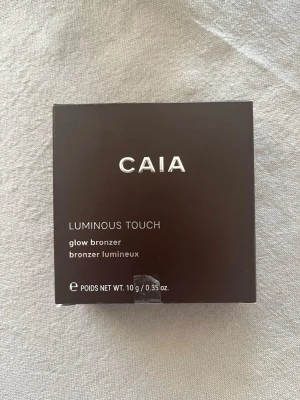 CAIA Luminous Touch Glow Bronzer - Säljer en CAIA Luminous Touch glow bronzer i färgen La Digue. Kommer i en stilren, mörkbrun fyrkantig förpackning med silvertext. Innehåller 10 g och ger ett snyggt glow till ansiktet. Perfekt för att skapa en solkysst look med naturlig lyster.
