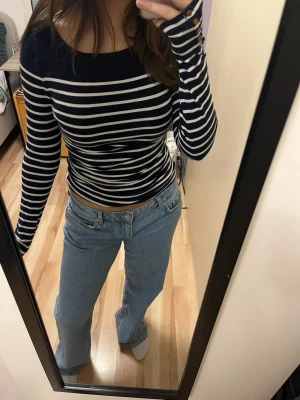 Marinblå och vit randig långärmad topp - Säljer en marinblå och vit långärmad topp med smala horisontella ränder. Toppen har en rundad hals och dekorativa knappar vid ärmsluten. Passformen är slim och materialet känns som mjuk bomull, perfekt för en chill och stilren look.