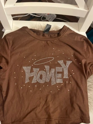 Brun croppad t-shirt med strass från SHEIN - Säljer en brun croppad t-shirt från SHEIN i storlek XS. T-shirten har korta ärmar och är gjord i mjuk bomull. Framsidan har texten 'HONEY' dekorerad med strass och ett glittrigt halomotiv ovanför, samt små strassdetaljer runtom. Perfekt för dig som gillar statement-plagg!