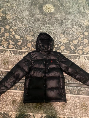 Svart pufferjacka från Polo Ralph Lauren - Säljer en svart pufferjacka från Polo Ralph Lauren med huva och dragkedja framtill. Jackan har en diskret röd logga på bröstet och två fickor med dragkedja. Perfekt för kalla dagar och riktigt snygg till streetwear-stilen.