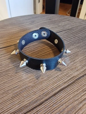Svart punk nitad läderchoker med spikes - Armband punk svart choker i läderimitation i PU med silvriga spikes och tryckknappar. 22 cm långt. Perfekt accessoar för dig som gillar edgy stil och vill sticka ut. Justerbar passform med två knappar. Snygg att matcha med streetwear eller rockiga outfits.