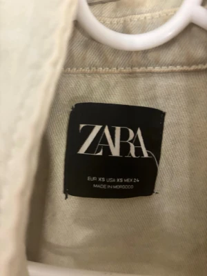 Beige jeansjacka från Zara XS - Snygg beige jeansjacka från Zara i storlek XS. Jackan har två stora bröstfickor med knappar och klassisk krage. Perfekt att slänga på sig för en chill och cool look. Materialet är robust och har en lite loose passform.