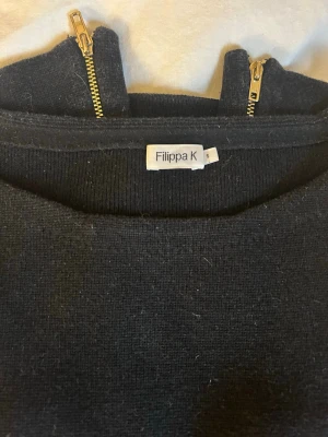 Svart tröja med dragkedjor från Filippa K - Snygg svart långärmad tröja från Filippa K med guldfärgade dragkedjor längs sidorna. Tröjan har en rund halsringning och är gjord i ett stickat material. Perfekt för dig som gillar stilrena detaljer och enkel design. Nypris över 2000kr, säljer för 300kr eller bud