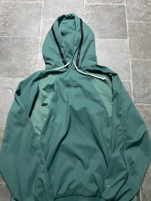 Grön vindjacka från Champion XL - Säljer en grön vindjacka från Champion i storlek XL. Jackan har huva med snörning, meshpaneler på sidorna och elastiska muddar. Champion-logga på bröstet och ärmen. Perfekt för dig som gillar sportig och avslappnad stil.