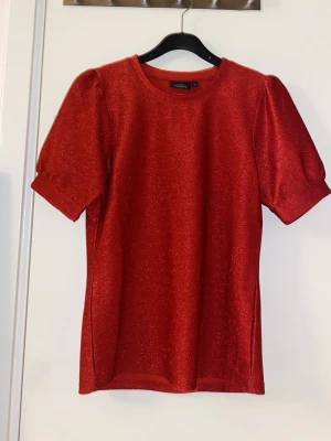 Röd glittrig topp från KappAhl XS - Säljer en röd glittrig topp från KappAhl i storlek XS. Toppen har puffiga korta ärmar och rund halsringning. Materialet är mjukt och skimrande, perfekt för att sticka ut. Enkel och snygg design som ger en cool vibe. Bara använd 2 gånger. 