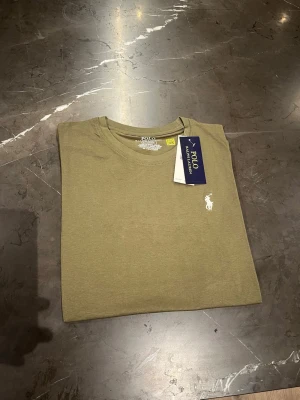 Olivgrön t-shirt från Polo Ralph Lauren - Snygg olivgrön t-shirt från Polo Ralph Lauren i storlek S. Klassisk rund hals och korta ärmar, med den ikoniska vita Polo-loggan broderad på bröstet. Tillverkad i mjuk bomull som känns skön mot huden. Perfekt för en chill och stilren look.