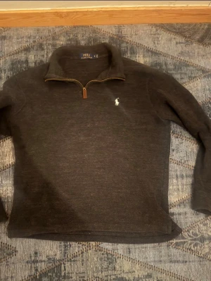 Mörkgrå half zip tröja Polo Ralph Lauren - Säljer en mörkgrå långärmad tröja från Polo Ralph Lauren i storlek S. Tröjan har en klassisk half zip med läderdetalj på dragkedjan och en vit broderad logga på bröstet. Materialet är mjukt och stickat, perfekt för kyliga dagar.