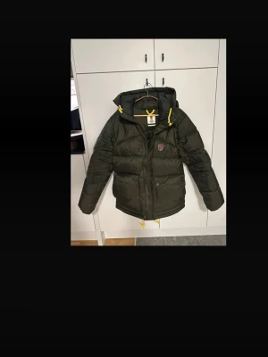 Grön Fjällräven Expedition dunjacka - Säljer en mörkgrön Fjällräven Expedition Series dunjacka med huva och gula detaljer. Jackan har puffad design, dragkedja och knappar framtill samt klassisk Fjällräven-logga på bröstet. Perfekt för kalla dagar och outdoor-äventyr. Finns på min v1nted för 6250