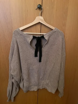 Beige stickad tröja med svart rosett i ryggen - Mysig beige stickad tröja med ribbad struktur och lång ärm. Tröjan har en snygg svart rosett bak som detalj och en relaxed, lite loose passform. Perfekt för lager på lager under höst och vinter. Har ingen lapp, men köpt på Chiquelle och var one size (passar en M).