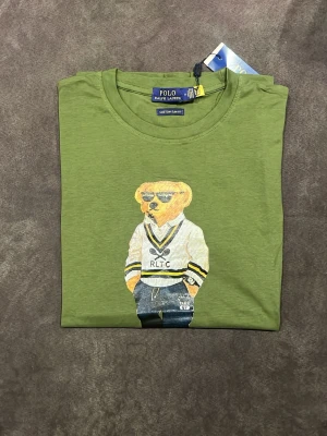 Grön Polo Bear t-shirt från Ralph Lauren - Snygg grön t-shirt från Ralph Lauren med Polo Bear-tryck på bröstet. T-shirten har rund halsringning och klassisk passform. Tillverkad i mjuk bomull och har en cool björn med solglasögon och RLTC-tröja på framsidan.
