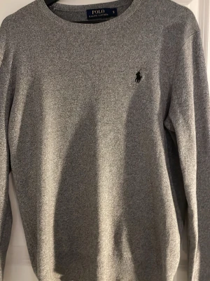 Grå stickad tröja från Polo Ralph Lauren - Snygg grå stickad tröja från Polo Ralph Lauren i storlek S. Klassisk rund hals och långärmad modell med den ikoniska broderade loggan på bröstet. Perfekt för dig som gillar stilrena och tidlösa plagg. Passar dig som vill ha en normal passform.