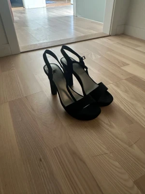 Svarta sandaler med klack - Säljer ett par svarta sandaler med hög klack och öppen tå. Skorna har en bred rem över tårna och en justerbar rem runt hälen. Materialet ser ut att vara mocka och sulan är platt framtill. Perfekt för dig som gillar stilrena och klassiska skor.