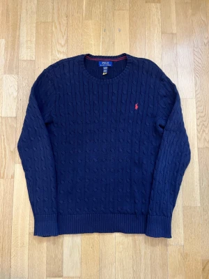 Marinblå stickad tröja Polo Ralph Lauren - Säljer en marinblå stickad tröja från Polo Ralph Lauren i storlek XL. Tröjan har ett klassiskt kabelstickat mönster och en rund hals. På bröstet finns den ikoniska röda Polo-loggan broderad. Perfekt för dig som gillar stilrena och tidlösa plagg.