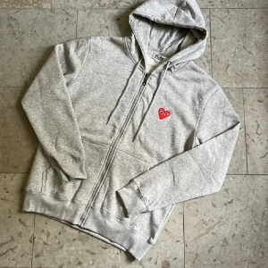 Grå Cdg play zip - Grå ziphoodie från Cdg Play med dragkedja, huva och snörning. På bröstet finns den ikoniska röda loggan. Tröjan är i storlek S och är i väldigt bra skick. Fraktas inom 24h✅