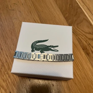 Silverfärgat armband från Lacoste - Stilrent armband från Lacoste i silverfärgad metall med rektangulära länkar och diskret Lacoste-logga på en av länkarna. Kommer i original ask och med vit tygpåse med dragsko, perfekt för att ge bort eller behålla själv. Passar killar tjejer pris kan diskuteras fraktar samma dag! 