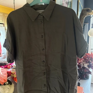 Olivgrön linneskjorta från Freequent - Olivgrön skjorta i linne från Freequent med klassisk krage, korta ärmar med knappdetalj.. Skjortan har knäppning framtill, är något längre baktill. Aldrig använd. (55% linne, 45% viskos)