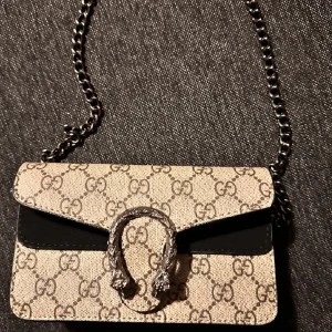 Gucci beige axelväska med kedja - Säljer en snygg beige axelväska från Gucci med klassiskt GG-mönster, silverfärgad kedja och ikoniskt spänne framtill. Väskan har svarta detaljer och är tillverkad i canvas med skinninslag. Perfekt för dig som vill sticka ut med en lyxig accessoar.