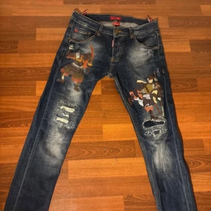 Dsquared2 jeans med samurai-tryck - Unika blå jeans från Dsquared2 med coola samurai-tryck på båda benen och slitna detaljer med hål och lagningar. Jeansen har klassisk femficksdesign, orange sömmar och en snygg tvättad finish. Perfekt för dig som vill sticka ut med din stil.
