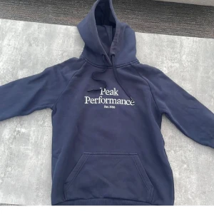 Mörkblå hoodie Peak Performance  - Snygg mörkblå hoodie från Peak Performance i storlek S. Tröjan har huva med snörning, stor magficka och vit logga på bröstet. Perfekt för chill eller träning, mjukt material och klassisk passform.