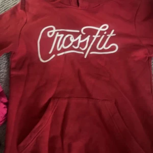 Röd CrossFit hoodie med huva - Röd hoodie från CrossFit med vit logga framtill. Klassisk känguruficka och huva. Perfekt för träning eller chill. Mjuk och skön känsla, passar dig som gillar sportig stil.