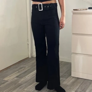 Svarta bootcut byxor med bälte - Svarta byxor med bootcut-modell och hög midja. Byxorna har ett brett bälte med stor silverfärgad spänne och hällor i midjan. Snygg passform som markerar midjan och ger en trendig siluett. Perfekta för dig som gillar en stilren och modern look.