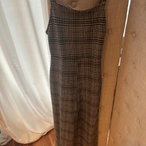 Rutig byxdress från Bershka, S - Snygg rutig byxdress från Bershka i storlek S. Byxdressen har smala justerbara axelband och vida ben, vilket ger en chill och trendig look. Färgen är en mix av beige, svart och brunt i ett klassiskt rutmönster. Perfekt för dig som gillar att sticka ut med mönster.