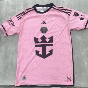 Inter Miami Messi 10 Adidas fotbollströja - Säljer en rosa Inter Miami fotbollströja från Adidas med Messi och nummer 10 på ryggen. Tröjan har svarta detaljer, klubbmärke på bröstet och stora tryck på både fram- och baksida. Tillverkad i lätt och ventilerande material, perfekt för fotboll eller supporterliv.
