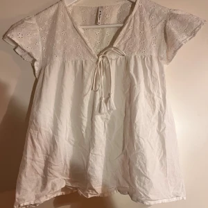 Vit blus med broderade detaljer - Superfin vit blus från Pepe Jeans med broderade blommor upptill och korta ärmar. Blusen har v-ringning med knytband och tofsar framtill. Luftig och lätt i bomull, perfekt för varma dagar. Söt och romantisk stil med volangliknande ärmar.