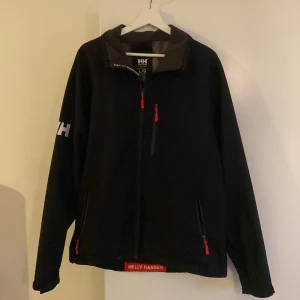 Svart Helly Hansen Jacka - Svart vindjacka från Helly Hansen med röda dragkedjor och HH-logga på ärmen. Jackan har ståkrage, hel dragkedja framtill och två fickor med dragkedja. Perfekt för blåsiga dagar och enkel att matcha med det mesta.