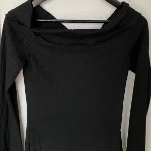 Svart långärmad topp från Dazy - Svart långärmad topp från Dazy med subtilt fiskbensmönster i tyget. Toppen har en bred, vikt krage och lätt utsvängda ärmar. Materialet är stretchigt och känns mjukt mot huden. Perfekt för dig som gillar stilrena och trendiga plagg.
