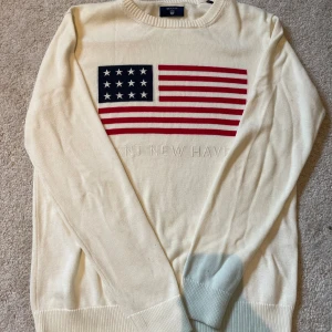 Vit GANT tröja med USA-flagga - Vit stickad tröja från GANT i 100% bomull med broderad amerikansk flagga och texten 'GANT NEW HAVEN' framtill. Klassisk rund halsringning och långa ärmar. Perfekt för dig som gillar preppy stil och vill sticka ut lite extra.