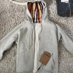 Grå hoodie från Burberry med dragkedja - Snygg grå hoodie från Burberry med klassiskt rutigt foder i huvan. Tröjan har dragkedja framtill, snörning i huvan och två fickor. Tillverkad i mjuk bomull, perfekt för chill dagar. Stilsäker detalj med Burberry-tag kvar.